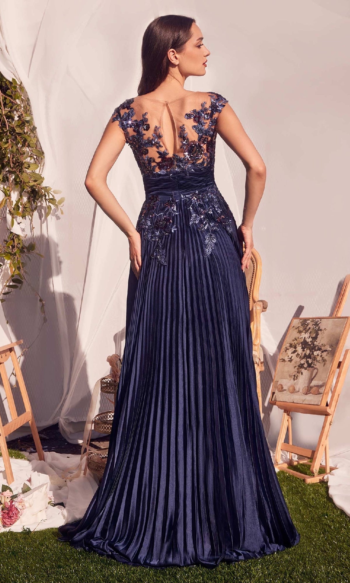 A woman stands outdoors in the Andrea & Leo A1355 Sequin Long Pleated Prom Dress, featuring a navy floor-length A-line skirt with sequin floral appliques on the sheer back and shoulders.